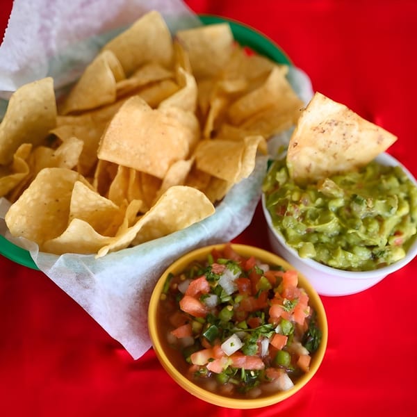 A basket of tortilla chips with bowls of guacamole and pico de gallo on a red table.