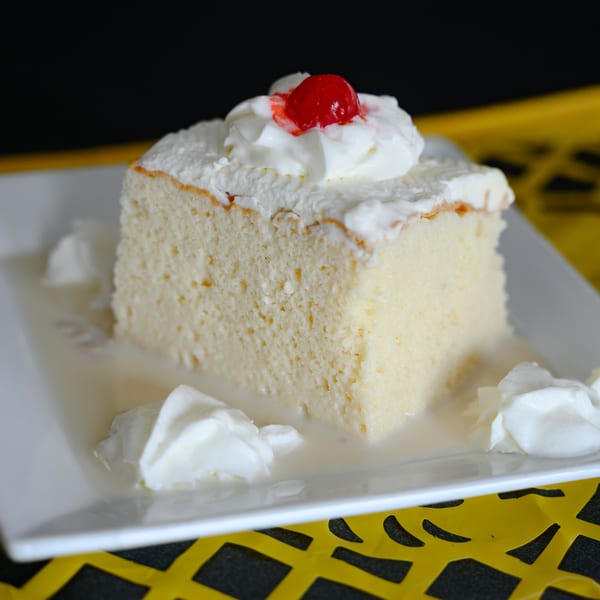 A slice of tres leches cake with whipped cream and a cherry on a white plate.